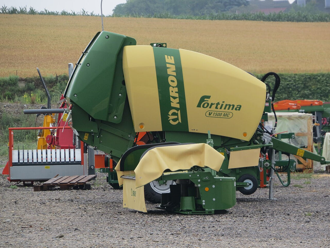 Krone Fortima V 1500 MC round baler parked on a gravel yard with a ripening grain field behind