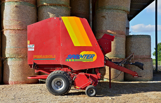 Red round baler parked in front of a tall stack of net-wrapped round bales in a shed
