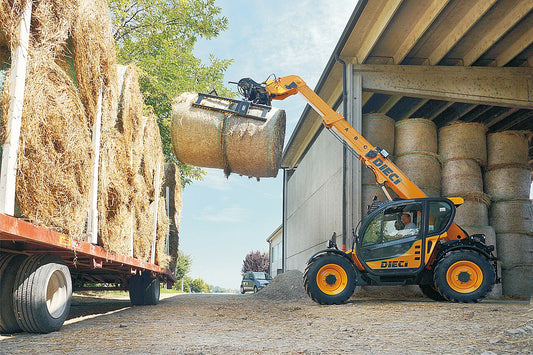 Telehandler with a round bale grab lifting a net-wrapped round bale off a flatbed trailer into a barn stacked with round bales