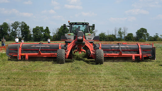 A Kuhn Merge Maxx 1090 twin-belt hay merger working behind a tractor, lifting two swaths onto its conveyor belts and laying them down as one clean windrow