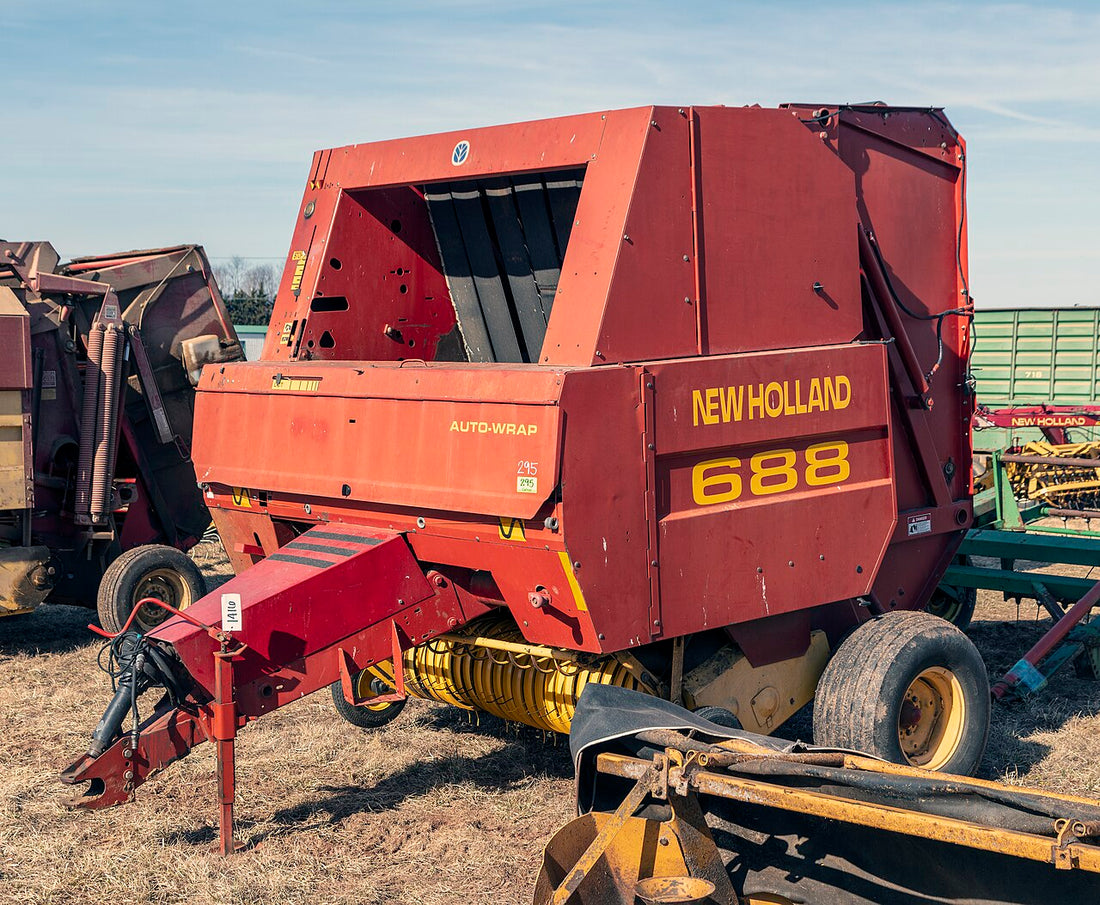 A New Holland 688 round baler parked outdoors on dormant grass during the off-season, tongue on its jack stand