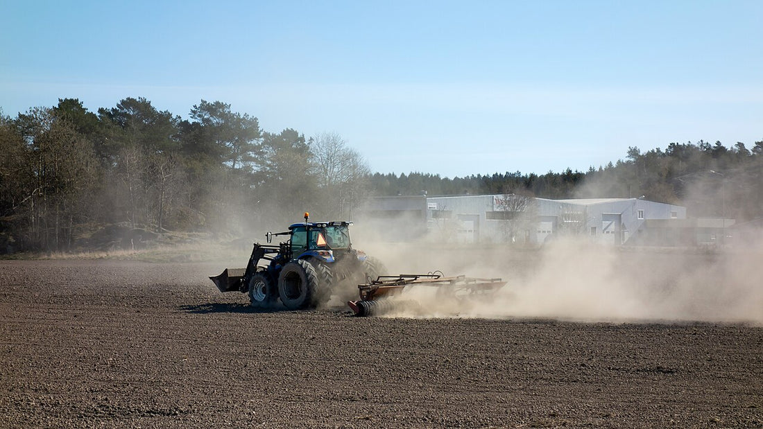 Tractor pulling a cultipacker across a worked field, raising a cloud of dust as it levels and firms the surface