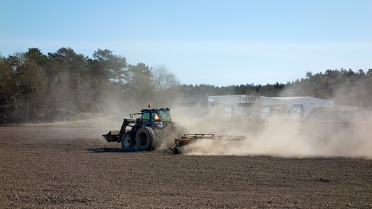Tractor pulling a cultipacker across a worked field, raising a cloud of dust as it levels and firms the surface