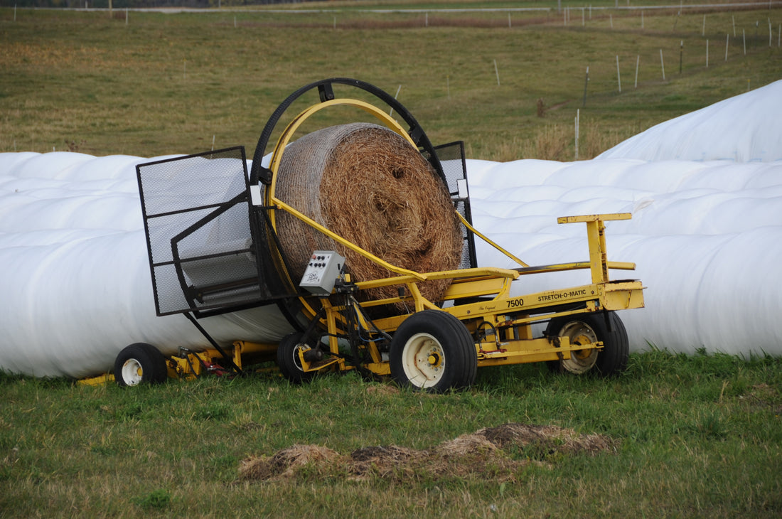 An AM Stretch-O-Matic 7500 inline tubular bale wrapper wrapping a net-wrapped round bale in white stretch film, with finished baleage tubes in the field behind