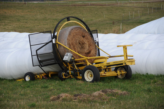 An AM Stretch-O-Matic 7500 inline tubular bale wrapper wrapping a net-wrapped round bale in white stretch film, with finished baleage tubes in the field behind