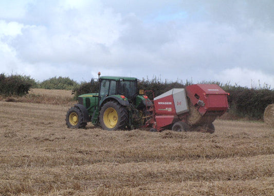 Green tractor pulling a red round baler through light straw windrows in a harvested wheat field
