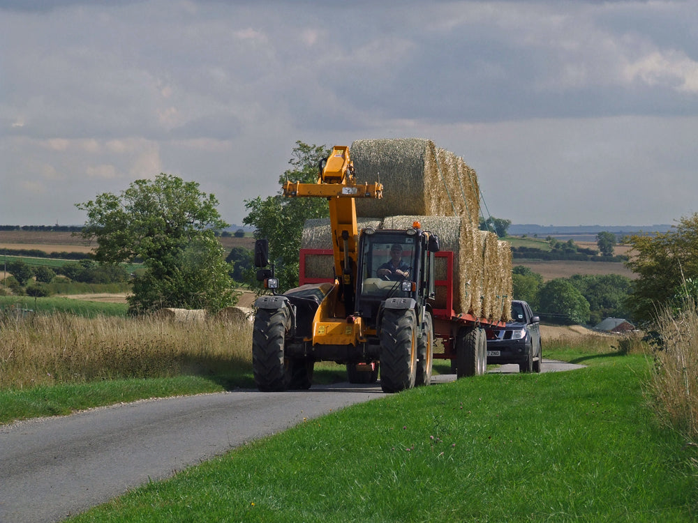 A telehandler towing a loaded bale trailer of round bales two high along a narrow country road, with a car following behind