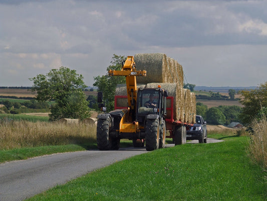 A telehandler towing a loaded bale trailer of round bales two high along a narrow country road, with a car following behind