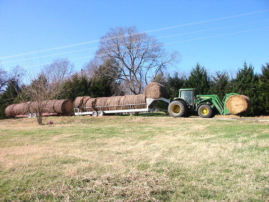 A rancher hauls round bales of hay on a flatbed trailer behind a tractor on a rural road in Grayson County, Texas