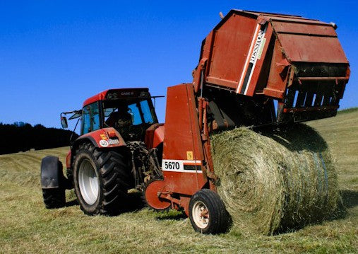 A Case IH tractor pulling a Hesston 5670 round baler that is discharging a finished round bale of dry hay in a sloping field