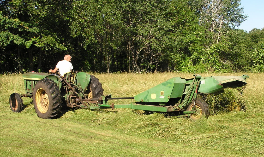 A John Deere tractor pulling a John Deere 1207 sickle-bar mower conditioner, laying a windrow of cut grass hay