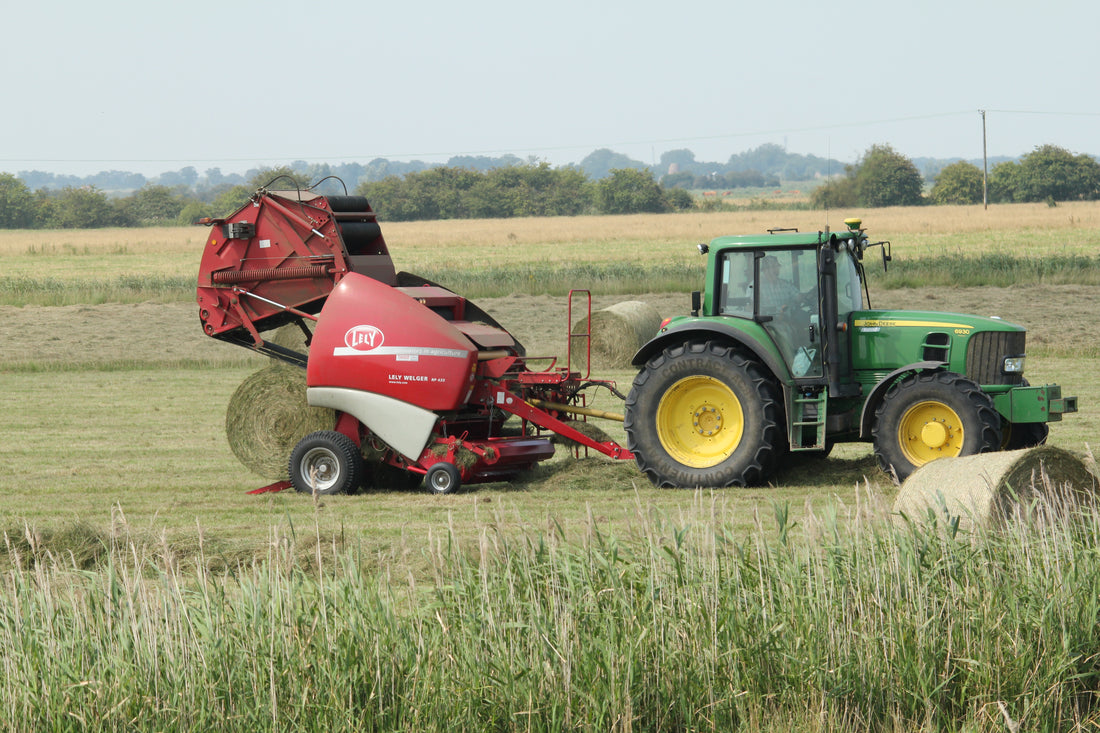 Red Lely Welger RP round baler towed by a green John Deere tractor baling hay in a mown field, with finished net-wrapped round bales behind