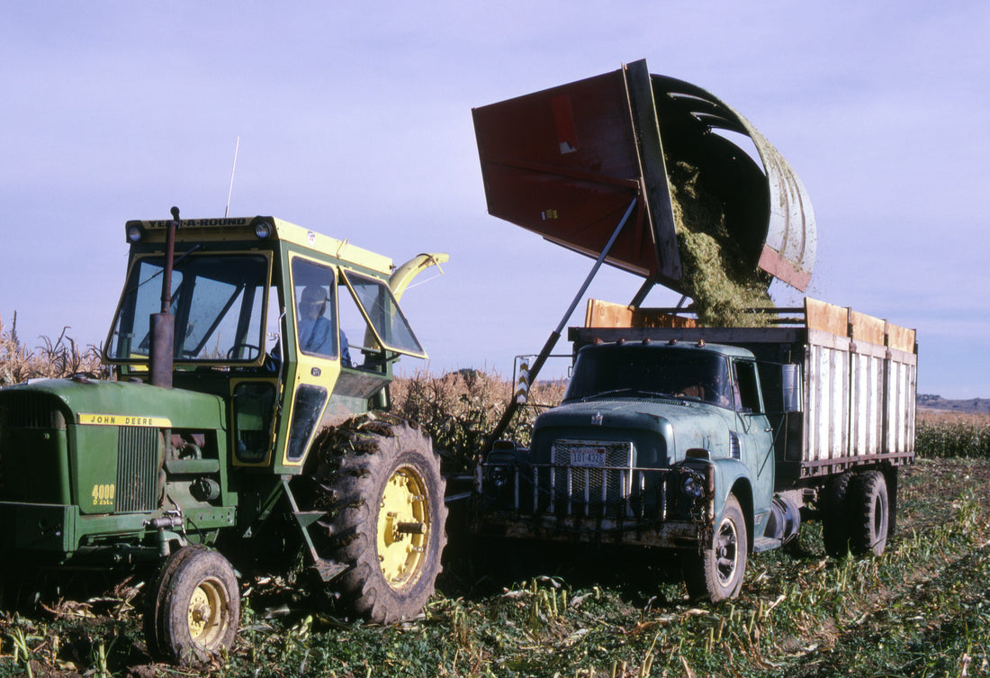 A John Deere tractor and a raised high-dump wagon tip freshly chopped corn silage into an old farm truck in a corn field near Joliet, Montana, in September 1978.