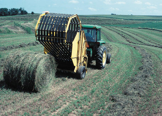 A round baler discharging a finished round hay bale with the belt-and-roller bale chamber visible, towed by a tractor in an Iowa hayfield